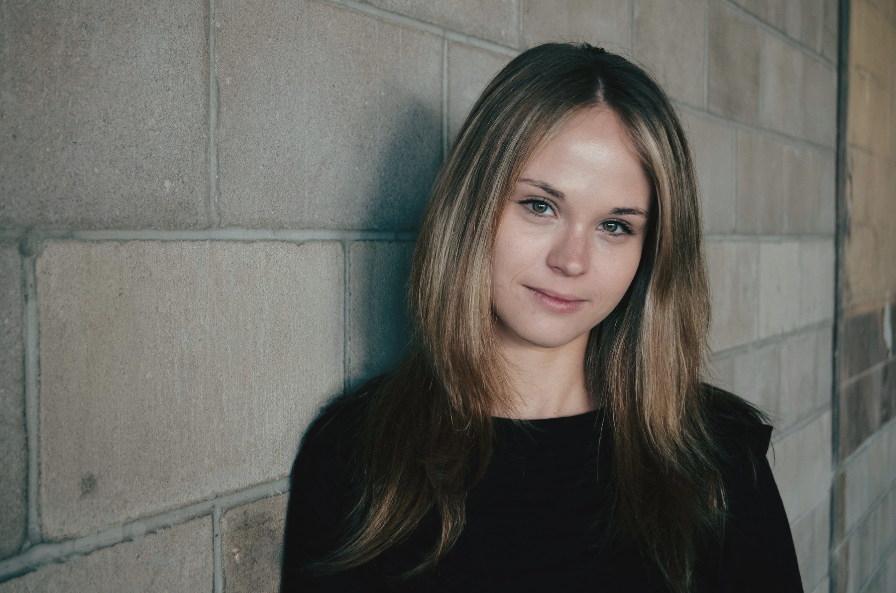 A woman in black leaning against a pale stone wall, looking into the lens