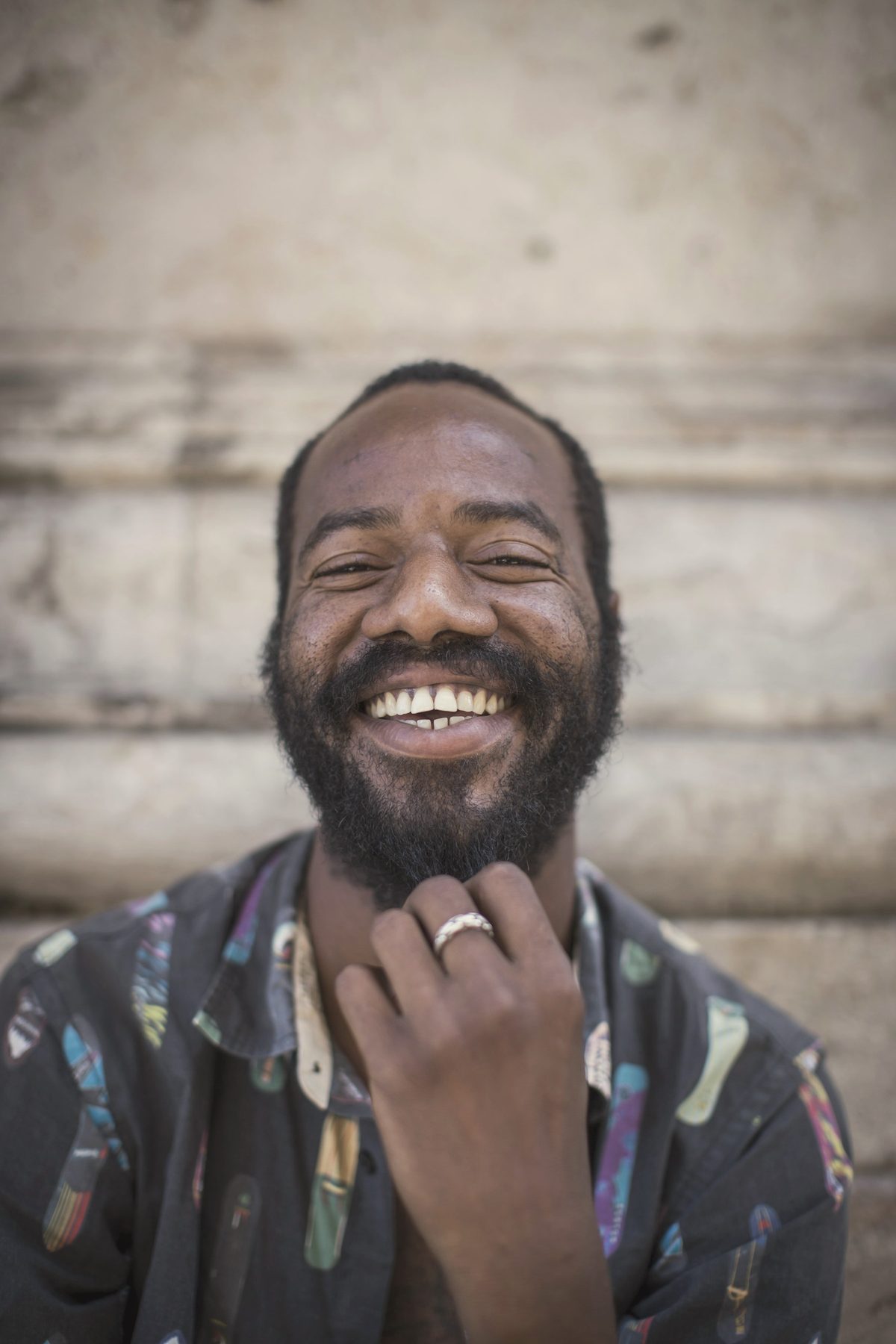 A man laughing with his hand at his beard, against a weathered wall