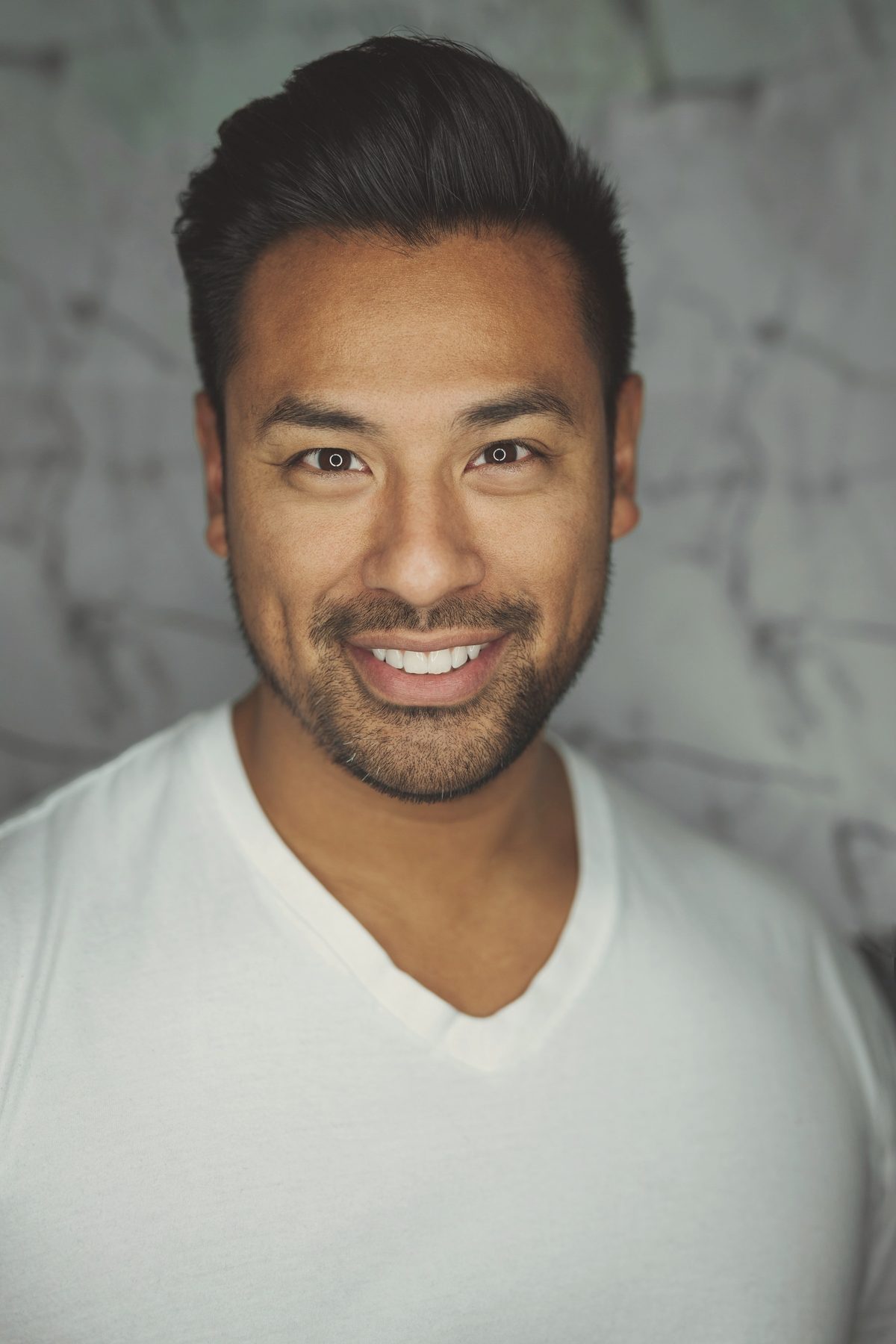 A man in a white T-shirt smiling, against a textured grey wall